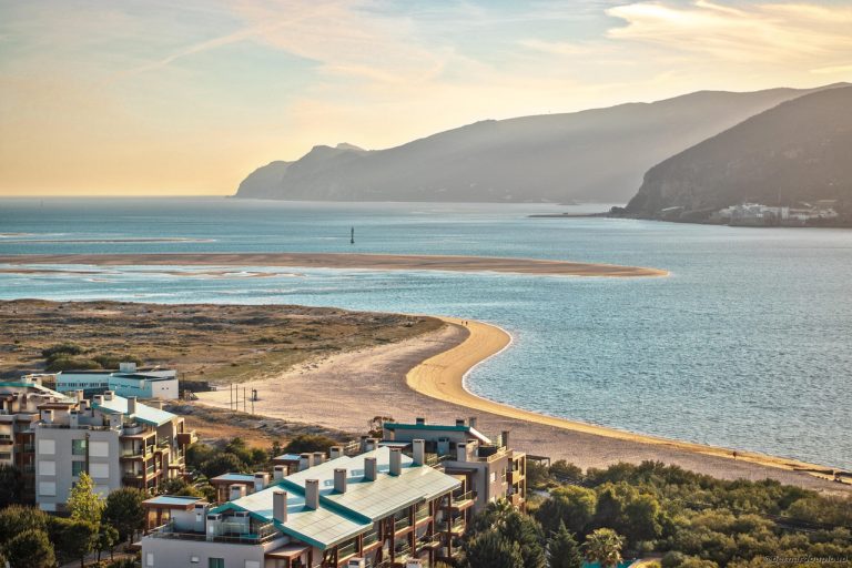 Plage de sable doré au Portugal, bordée d’eaux bleues, avec des immeubles au premier plan et des collines à l’horizon