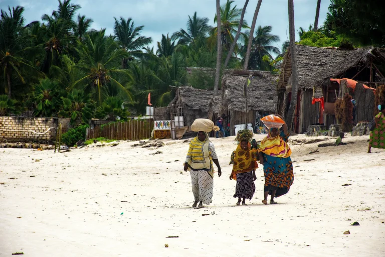 Trois personnes en vêtements colorés marchant sur une plage bordée de palmiers et d'habitations traditionnelles.